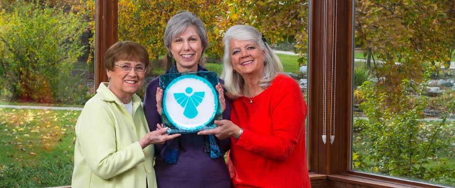 three elderly women holding a blue plate with an angel with a fall colored trees in the background at Attic Angel Community